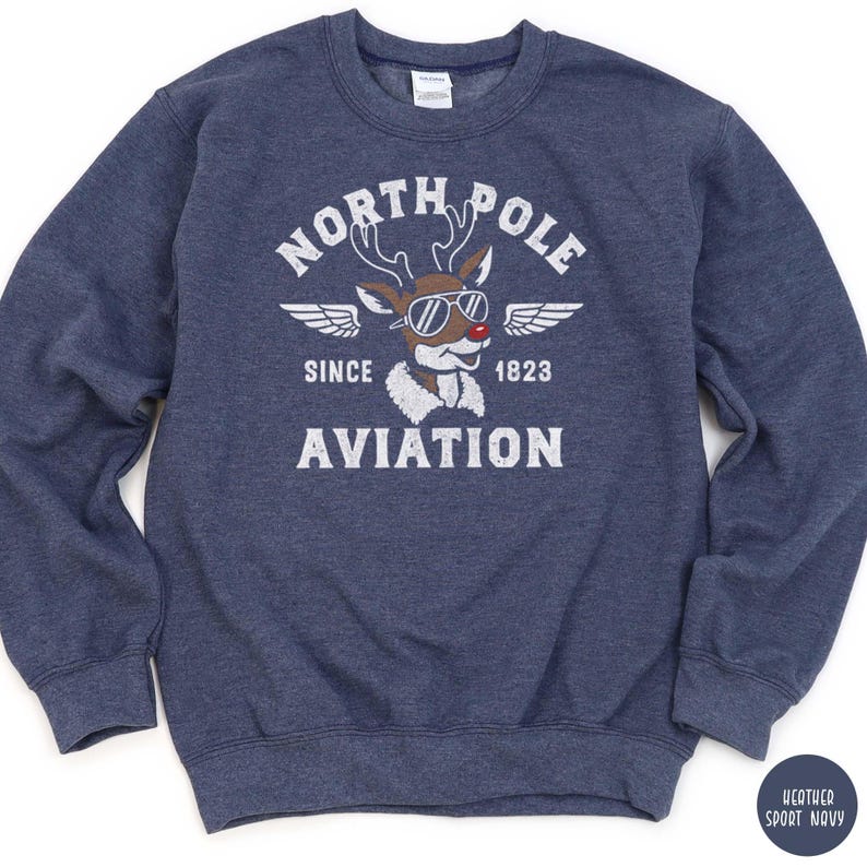 May include: A navy blue sweatshirt with a graphic featuring a reindeer wearing sunglasses and the words "NORTH POLE AVIATION SINCE 1823." The design includes wings and a vintage aesthetic. The sweatshirt is a heather sport navy color.