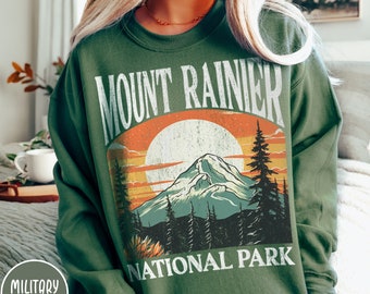 May include: A green sweatshirt with a vintage-style graphic of Mount Rainier National Park. The graphic features a mountain range, a sunset, and the words "Mount Rainier National Park".
