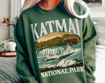 May include: Green sweatshirt with a vintage-style graphic of Katmai National Park. The graphic features a brown bear standing on a waterfall with salmon swimming around it.