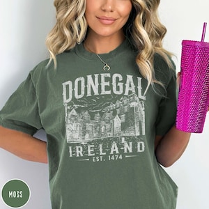 May include: Moss green t-shirt with a distressed white graphic of "Donegal Ireland" and a castle illustration. The text "Est. 1474" is below the graphic. A pink studded tumbler with a straw is held in the hand.