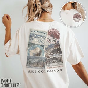 May include: Ivory-colored t-shirt featuring a collage of ski resort imagery from Colorado, including Vail, Aspen, and Telluride. The design incorporates vintage-style stamps and the text "SKI COLORADO". The shirt is made of Comfort Colors material.