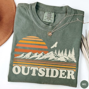 May include: Moss green t-shirt with a retro sunset and mountain graphic. The design features an orange and yellow striped sun, white mountains, and the word "OUTSIDER" in a bold font. A silver necklace with a pendant is draped on the shirt.