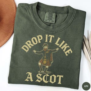 Puede incluir: Camiseta verde oliva con el texto "DROP IT LIKE A SCOT" en una fuente vintage. El gr&aacute;fico presenta una ilustraci&oacute;n de dibujos animados de un hombre con vestimenta escocesa tradicional, incluyendo un kilt y un sombrero. La camiseta tiene cuello redondo y mangas cortas.