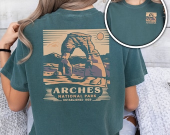 Op de afbeelding: Een tealblauw t-shirt met een vintage-stijl illustratie van Delicate Arch in Arches National Park, Utah. De tekst "Arches National Park Established 1929" is onder de illustratie gedrukt.