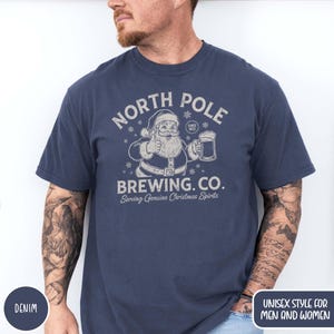 May include: A denim blue t-shirt with a graphic of Santa Claus giving a thumbs up and holding a beer mug. The text reads "NORTH POLE BREWING CO. Serving Genuine Christmas Spirits". The shirt is a unisex style.