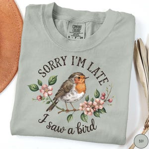May include: A light green t-shirt with a robin perched on a branch with pink flowers. The text "Sorry I'm Late I saw a bird" is printed on the shirt.