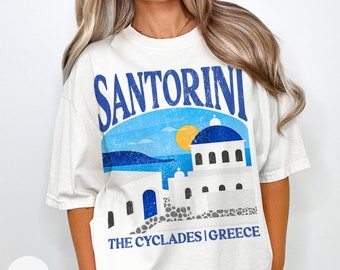 May include: A white t-shirt with a blue and white graphic of a building in Santorini, Greece. The text "Santorini" is in blue and the text "The Cyclades | Greece" is in black.