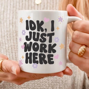 May include: White ceramic mug with the text "IDK, I JUST WORK HERE" in black, decorated with pastel hearts, stars, and flowers. The mug has a white handle and is held by a person.