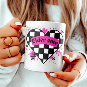 May include: White ceramic mug with a black and white checkered heart design. The heart has a pink banner with the text "Elder emo" on it. The mug has pink and black splatters around the heart.