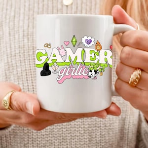 Può includere: Tazza in ceramica bianca con un design verde, rosa e nero che dice "gamer girlie". Il design include cuori, un diamante, una nuvola, un coniglio, un anello, un fuoco, una pianta e una mucca.