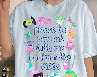 Puede incluir: Una camiseta azul claro con el texto please be patient with me im from the 1900s rodeado de coloridas ilustraciones de objetos de los años 90, incluyendo un Furby, un disquete, una consola de juegos portátil, un chupete y un reproductor de CD.