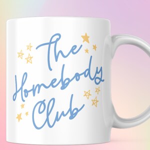 Pode incluir: Caneca de cerâmica branca com uma inscrição azul que diz "The Homebody Club" e estrelas douradas ao redor do texto.
