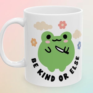 May include: White ceramic mug with a green frog holding a knife and the text "Be Kind Or Else" in black lettering. The frog is surrounded by small flowers and clouds.