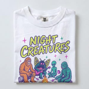 Può includere: Una t-shirt bianca con una stampa grafica che riporta il testo "NIGHT CREATURES" in verde lime. L'illustrazione raffigura un sasquatch marrone, una creatura simile a una falena che suona la chitarra, un alieno verde e un mostro della palude.