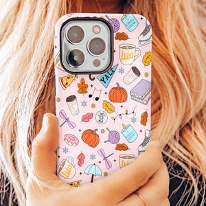 Op de afbeelding: Een roze telefoonhoesje met een herhaald patroon van herfstthema-illustraties, waaronder pompoenen, bladeren, koffiemokken en een boek. De hoes bevat ook het woord "Yale" en de zin "I smell snow".