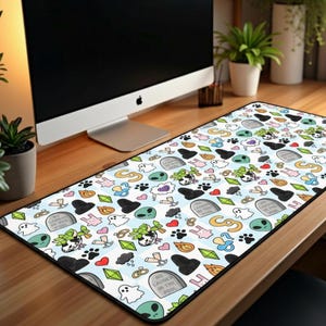 May include: A rectangular desk mat featuring a white background with a repeating pattern of cartoon ghosts, aliens, tombstones, paw prints, and hearts. The mat is designed to protect the desk surface and provide a smooth surface for a mouse.