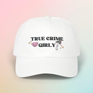 Könnte beinhalten: Weiße Baseballkappe mit schwarzem gesticktem Text "TRUE CRIME GIRLY" mit einem rosa Herzen und einem schwarzen Pilz-Motiv.
