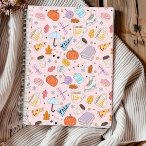 Puede incluir: Un cuaderno con espiral y cubierta rosa con un patrón repetido de calabazas, rosquillas, tazas de café y otras ilustraciones otoñales. Encuadernación en espiral plateada.