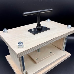 May include: A wooden press with a metal handle and a threaded rod. The press is made of light wood and has a rectangular shape. The handle is black and has a T-shaped grip. The threaded rod is silver and is attached to the handle. The press is used for pressing objects, such as fruits or vegetables, to extract juice or oil.