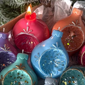 May include: A box of colorful, decorative candles shaped like ornaments. The candles are in shades of red, purple, blue, teal, orange, and gray. One candle is lit, and the box is lined with white paper. The candles have glitter accents.