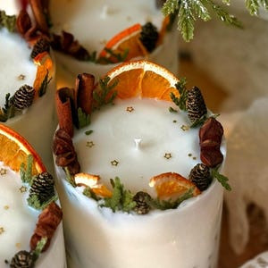 May include: White pillar candles decorated with dried orange slices, pine cones, cinnamon sticks, and sprigs of greenery. The candles have a scattering of gold star-shaped embellishments. The candles are a festive, decorative item.
