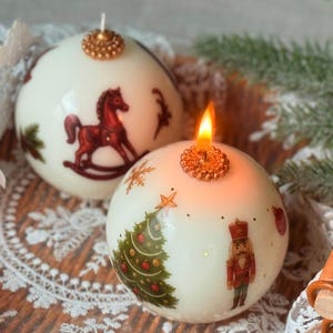May include: Two round, white Christmas candles with festive illustrations. One candle features a rocking horse, the other a Christmas tree and a nutcracker. Both have a gold-colored decorative top and are lit with a warm glow.
