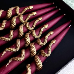 Puede incluir: Un conjunto de velas cónicas de color burdeos intenso, cada una con una serpiente dorada texturizada enroscada a lo largo de la vela. Las serpientes tienen escamas detalladas y están dispuestas en varios patrones sinuosos sobre la cera.