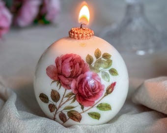 May include: A spherical white candle decorated with red roses and green leaves. The candle has a lit wick and a textured, decorative top. The candle is placed on a soft, textured surface.