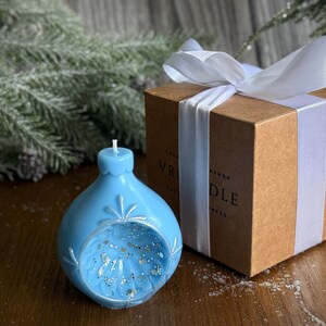 May include: A light blue, ornament-shaped candle with silver glitter and snowflake details. The candle sits next to a brown gift box tied with a white ribbon. The box has the text "VR CANDLE WORKSHOP".