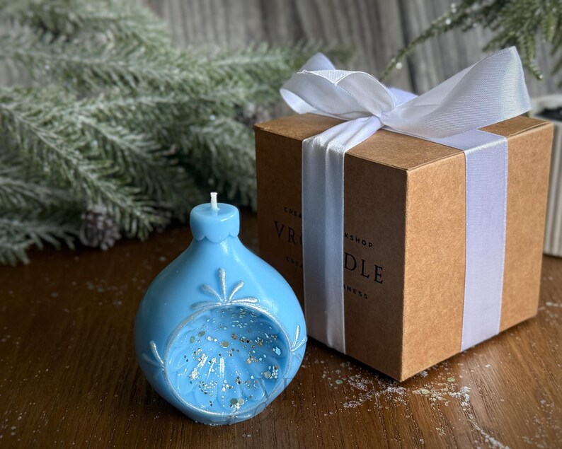 May include: A light blue, ornament-shaped candle with silver glitter and snowflake details. The candle sits next to a brown gift box tied with a white ribbon. The box has the text "VR CANDLE WORKSHOP".