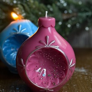 May include: A close-up of two decorative, spherical candles. The front candle is a vibrant pink with silver accents and glitter inside. The second candle is blue. Both candles have a floral design and a wick.