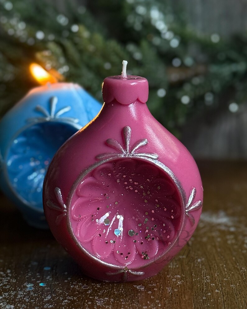May include: A close-up of two decorative, spherical candles. The front candle is a vibrant pink with silver accents and glitter inside. The second candle is blue. Both candles have a floral design and a wick.