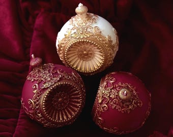 Puede incluir: Tres adornos esféricos ornamentados con intrincados diseños de filigrana dorada. Uno es blanco y dorado, mientras que los otros dos son de un borgoña profundo con detalles dorados. Los adornos descansan sobre una rica superficie de terciopelo rojo.