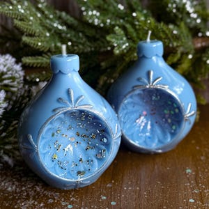 May include: Two light blue, teardrop-shaped candles with silver accents and glitter inside. The candles have a decorative floral design around the opening and a small wick at the top. The background includes evergreen branches.