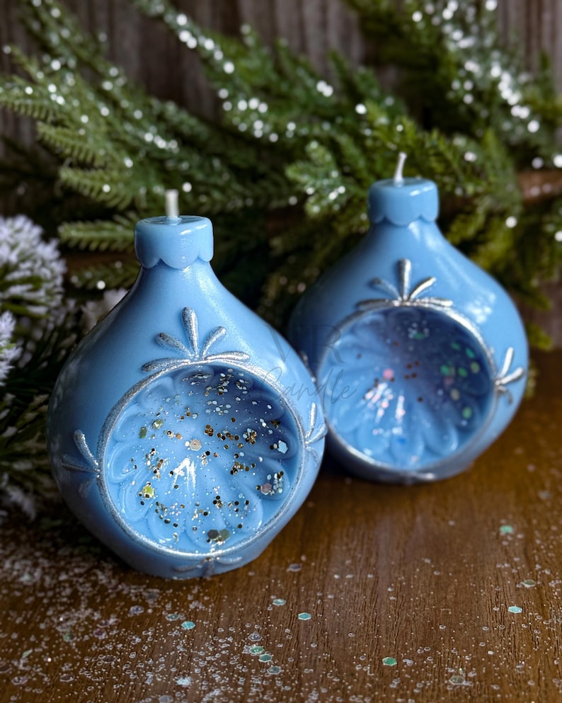 May include: Two light blue, teardrop-shaped candles with silver accents and glitter inside. The candles have a decorative floral design around the opening and a small wick at the top. The background includes evergreen branches.