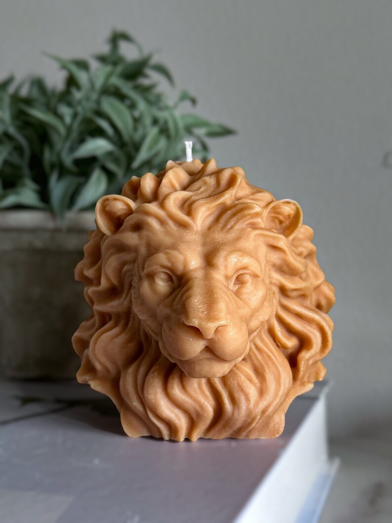 May include: A detailed lion head candle made of beige wax. The candle has a realistic design with a mane and a serious expression.