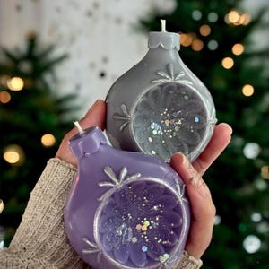 May include: Two Christmas ornament-shaped candles, one gray and one purple, are held in a person's hands. The candles have a floral design with glitter inside. The background is blurred with a Christmas tree and bokeh lights.
