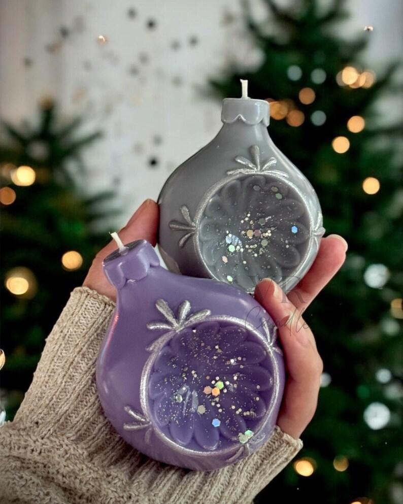 May include: Two Christmas ornament-shaped candles, one gray and one purple, are held in a person's hands. The candles have a floral design with glitter inside. The background is blurred with a Christmas tree and bokeh lights.