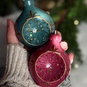 May include: Two decorative, teardrop-shaped candles in teal and magenta. Each candle features a gold-colored circular design with glitter accents. The candles are designed to resemble Christmas ornaments. The candles are handmade.