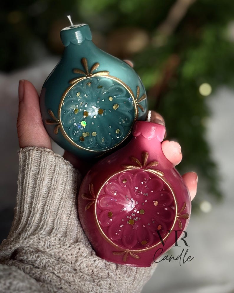 May include: Two decorative, teardrop-shaped candles in teal and magenta. Each candle features a gold-colored circular design with glitter accents. The candles are designed to resemble Christmas ornaments. The candles are handmade.