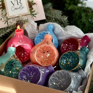May include: A box of colorful, decorative candles in various shapes and colors, including pink, orange, teal, and purple. Some candles have glitter accents and are shaped like ornaments. The box also contains a card with the text "VR Candle".