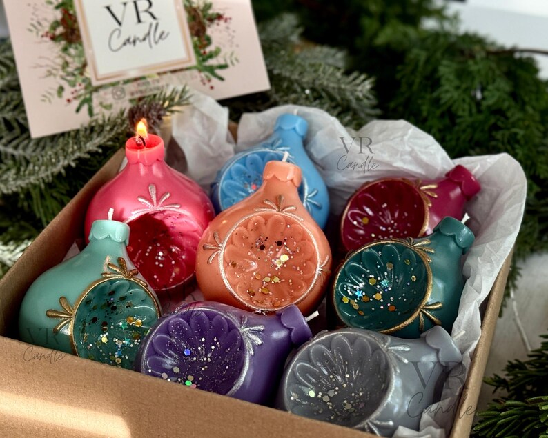 May include: A box of colorful, decorative candles in various shapes and colors, including pink, orange, teal, and purple. Some candles have glitter accents and are shaped like ornaments. The box also contains a card with the text "VR Candle".