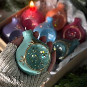 May include: A hand holds a teal-colored candle shaped like an ornament, with a gold-colored trim and a clear center filled with glitter. Other colorful candles are in the background. The candles are in a cardboard box. The text "VR Candle" is visible.