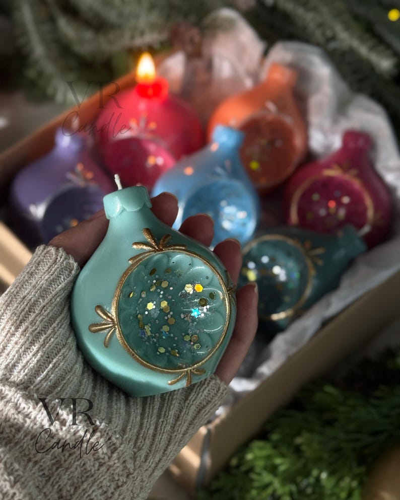 May include: A hand holds a teal-colored candle shaped like an ornament, with a gold-colored trim and a clear center filled with glitter. Other colorful candles are in the background. The candles are in a cardboard box. The text "VR Candle" is visible.
