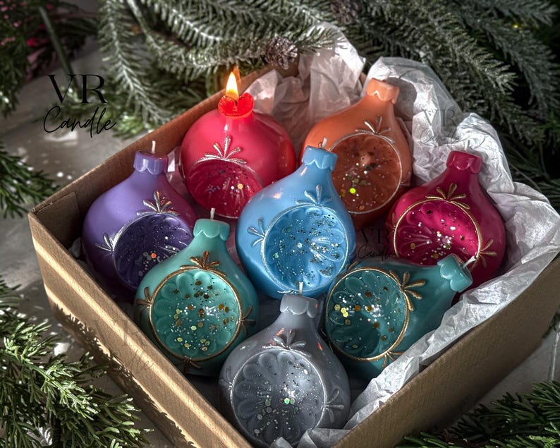 May include: A box of colorful, decorative candles shaped like ornaments. The candles are in shades of red, purple, blue, teal, orange, and gray. One candle is lit, and the box is lined with white paper. The candles have glitter accents.