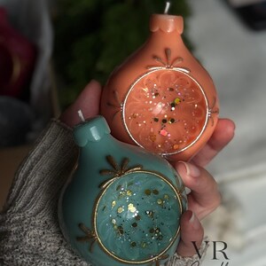 May include: Two decorative, teardrop-shaped candles. One is coral-colored, the other teal. Both have a circular, open design with glitter and gold accents. The candles are from VR Candle.