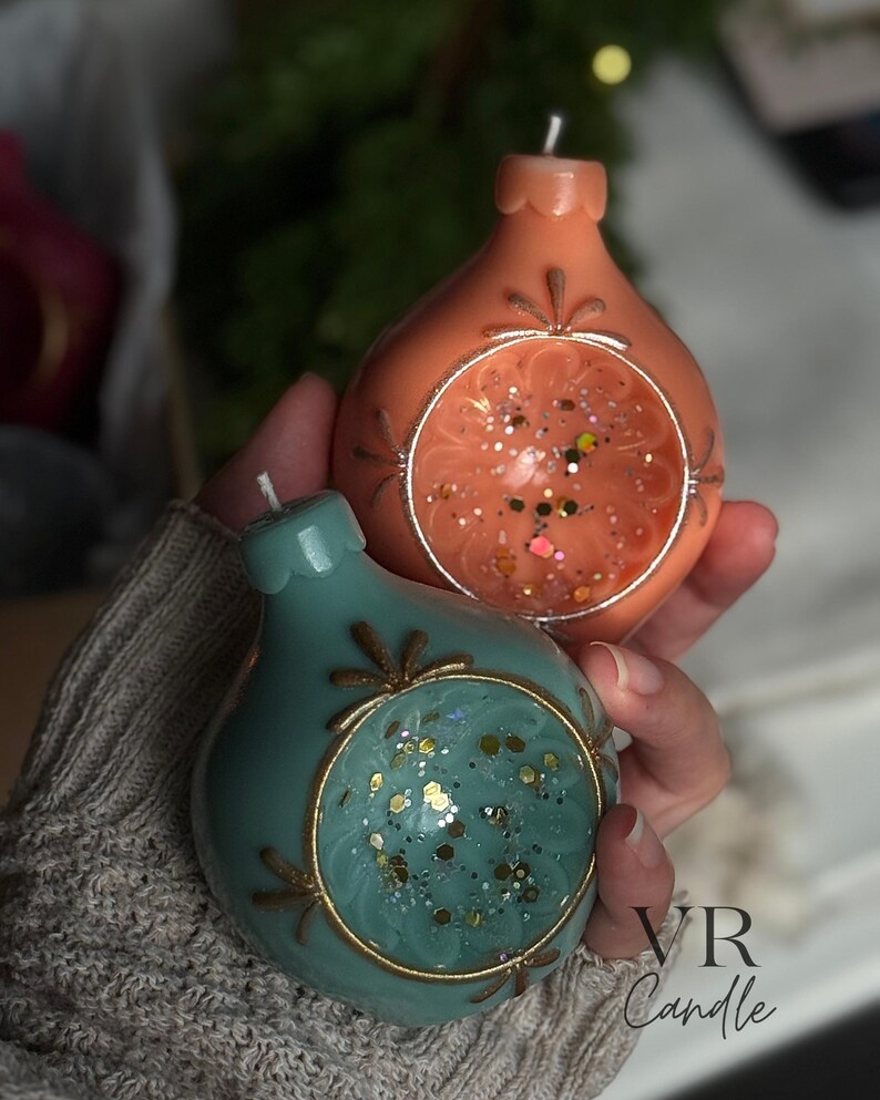 May include: Two decorative, teardrop-shaped candles. One is coral-colored, the other teal. Both have a circular, open design with glitter and gold accents. The candles are from VR Candle.
