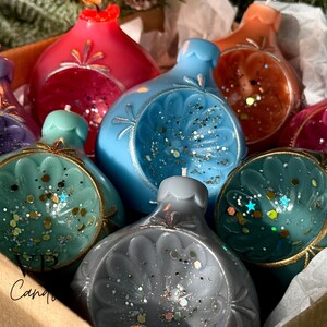 May include: A collection of colorful, decorative candles shaped like ornaments. The candles are in shades of red, blue, teal, purple, orange, and gray, with gold accents and glitter. They are arranged in a cardboard box.