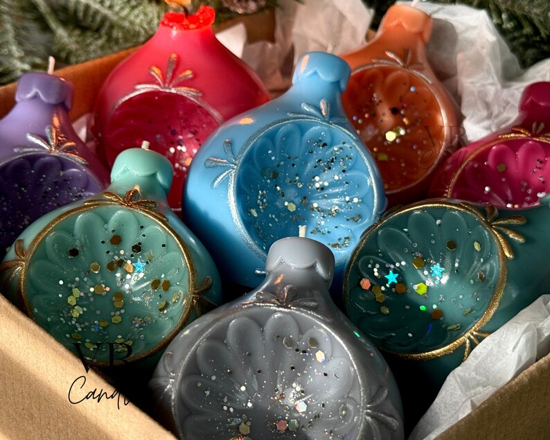 May include: A collection of colorful, decorative candles shaped like ornaments. The candles are in shades of red, blue, teal, purple, orange, and gray, with gold accents and glitter. They are arranged in a cardboard box.