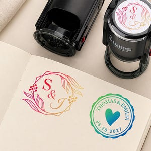May include: Two black self-inking stamps positioned above an open notebook. One stamp impression features a floral frame with the letters S&J in a gradient red to yellow. The other impression is a circular seal with the text THOMAS & EMILIA, a heart, and the date 20.05.2021 in a gradient green to blue.